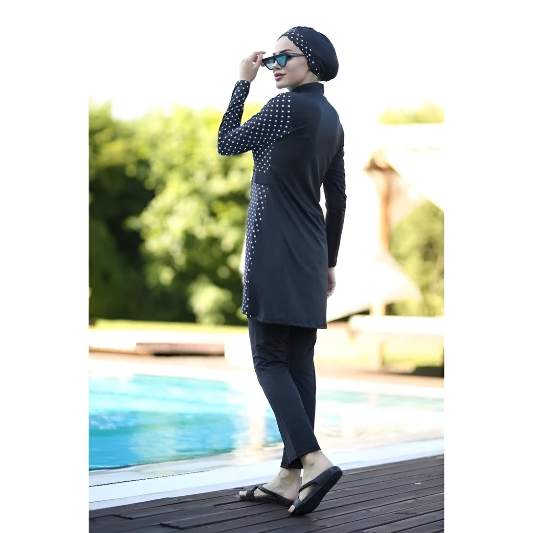Une femme portant un maillot de bain musulman noir à pois blancs se tient au bord d'une piscine. Elle porte également un foulard noir et des lunettes de soleil.