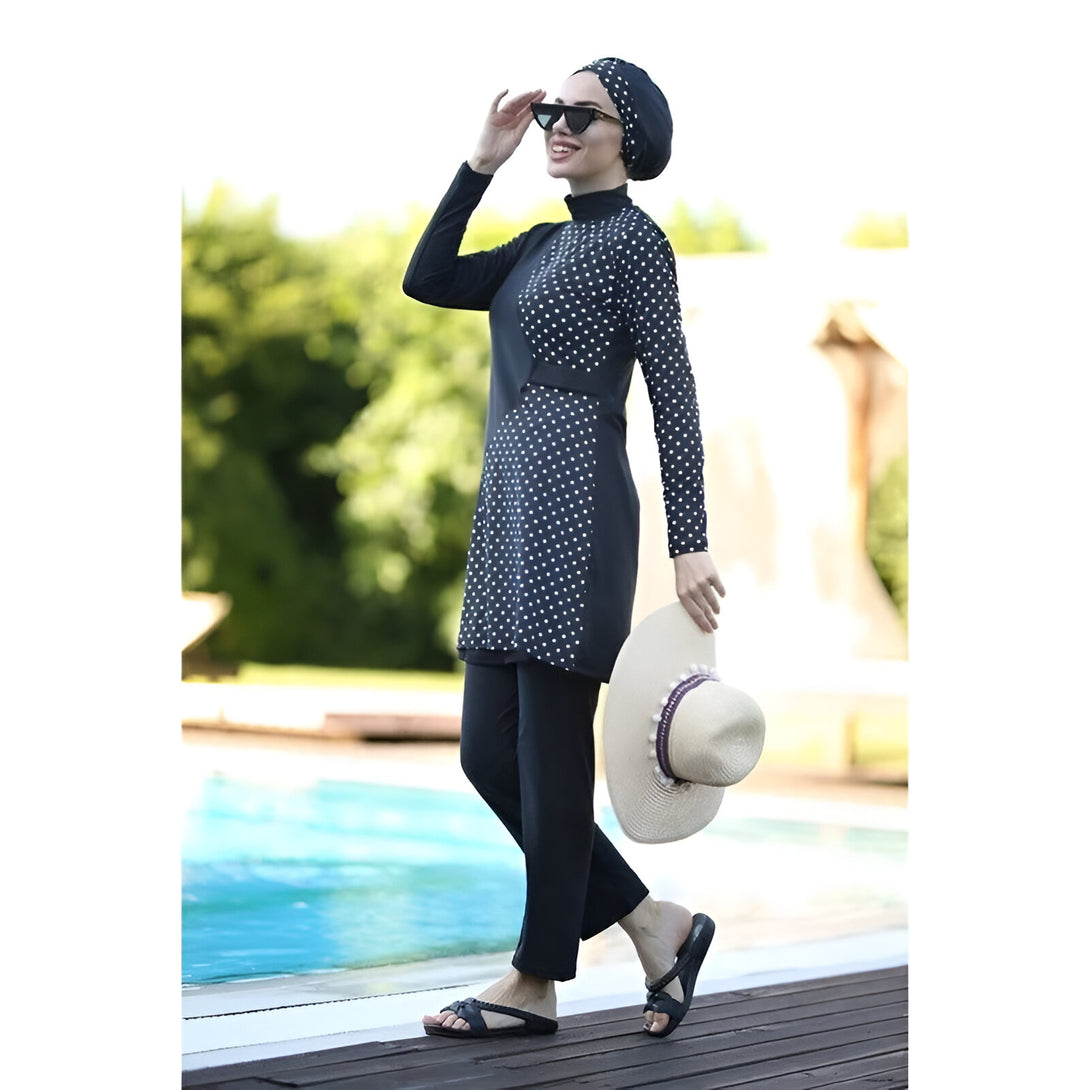 Une femme portant un maillot de bain musulman noir à pois blancs, un hijab et des lunettes de soleil, se tient près d'une piscine.  Elle tient un chapeau de paille dans sa main.