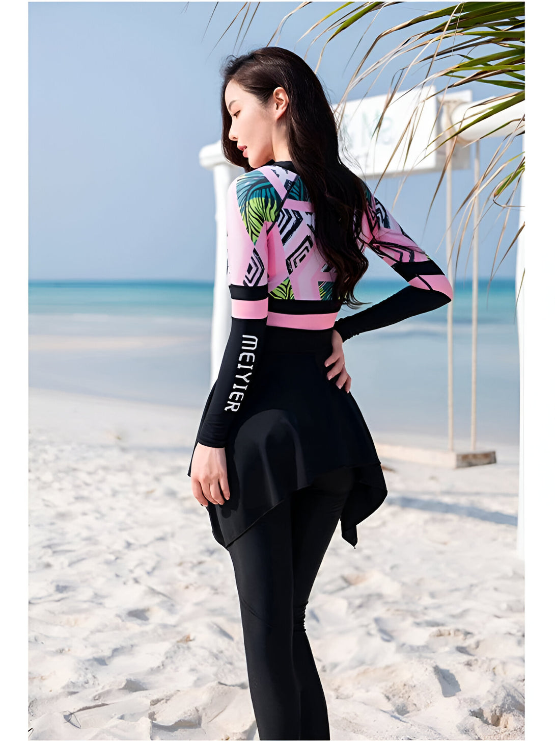 Une femme porte un maillot de bain musulman à manches longues rose et noir avec un imprimé tropical. Le maillot de bain est composé d'un haut à manches longues et d'un pantalon long. Elle se trouve sur une plage de sable blanc.