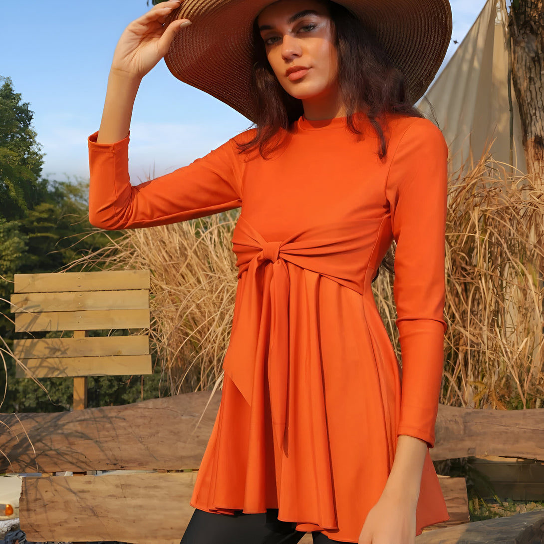 Une femme portant une robe de plage musulmane orange avec un nœud à la taille, sous un chapeau de paille, dans un paysage naturel.