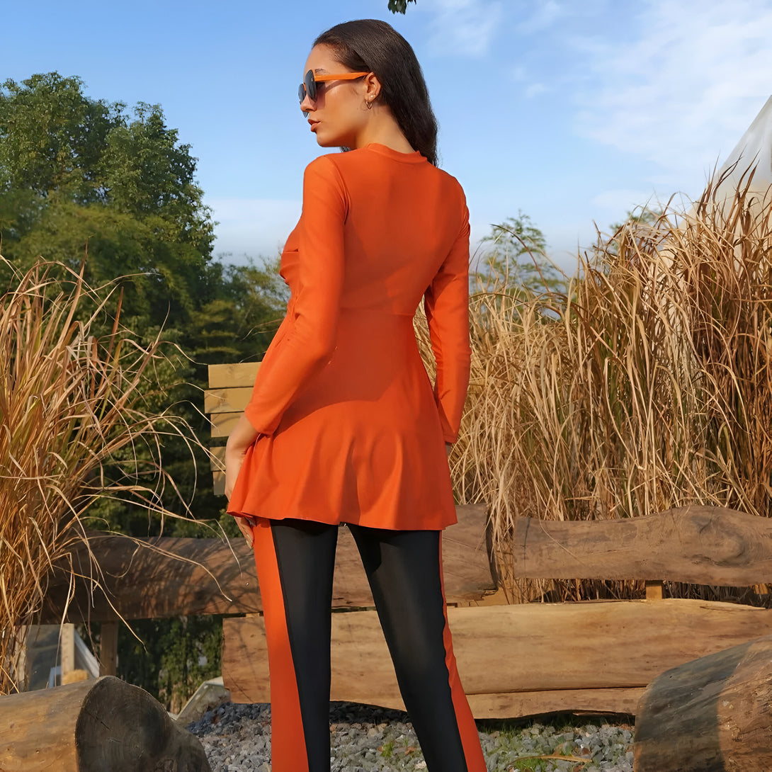 Une femme portant un maillot de bain musulman orange à manches longues et un pantalon noir, debout devant un banc en bois.