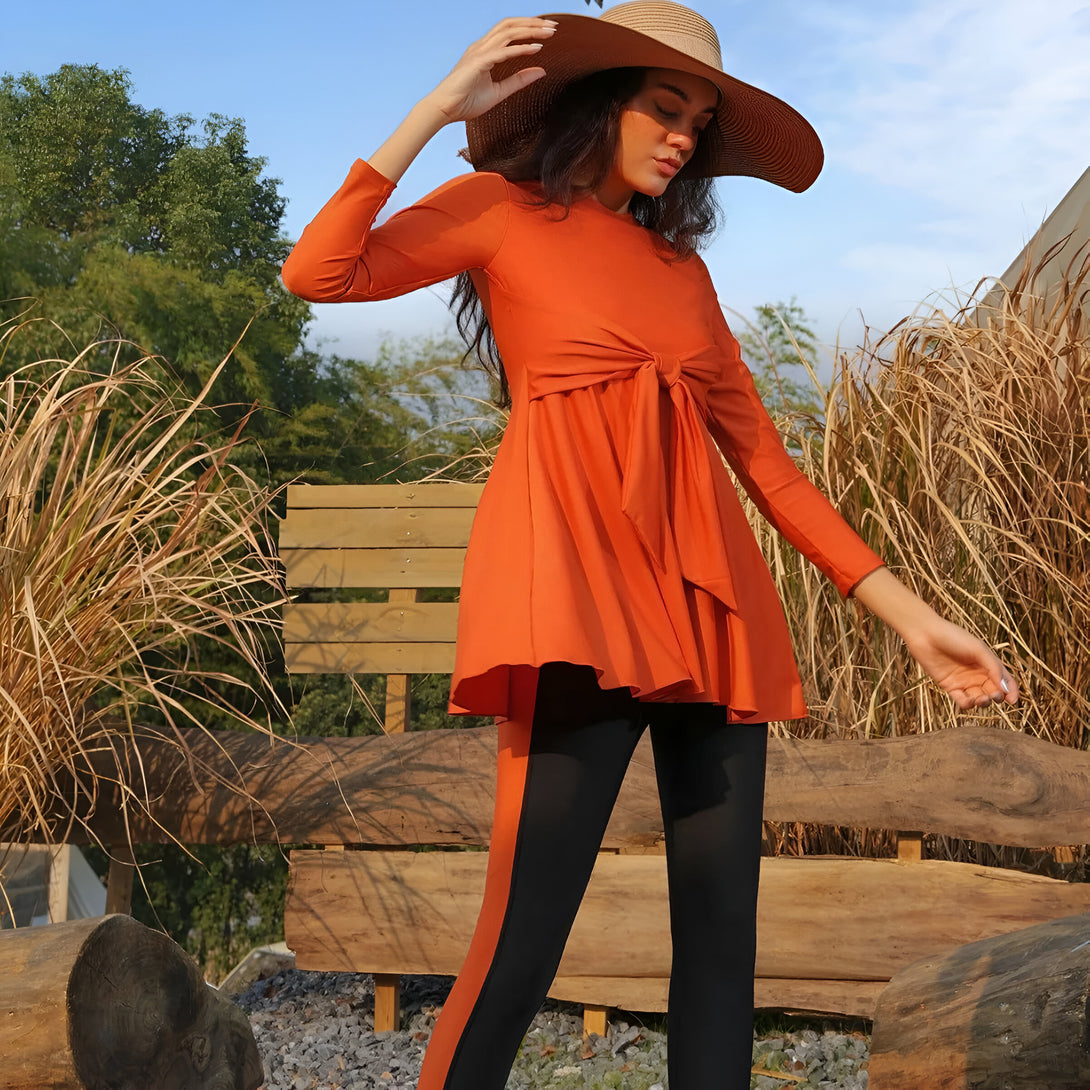 Une femme portant un maillot de bain musulman orange avec un nœud à la taille et un pantalon noir. Elle porte un chapeau de paille et pose près d'un banc en bois.