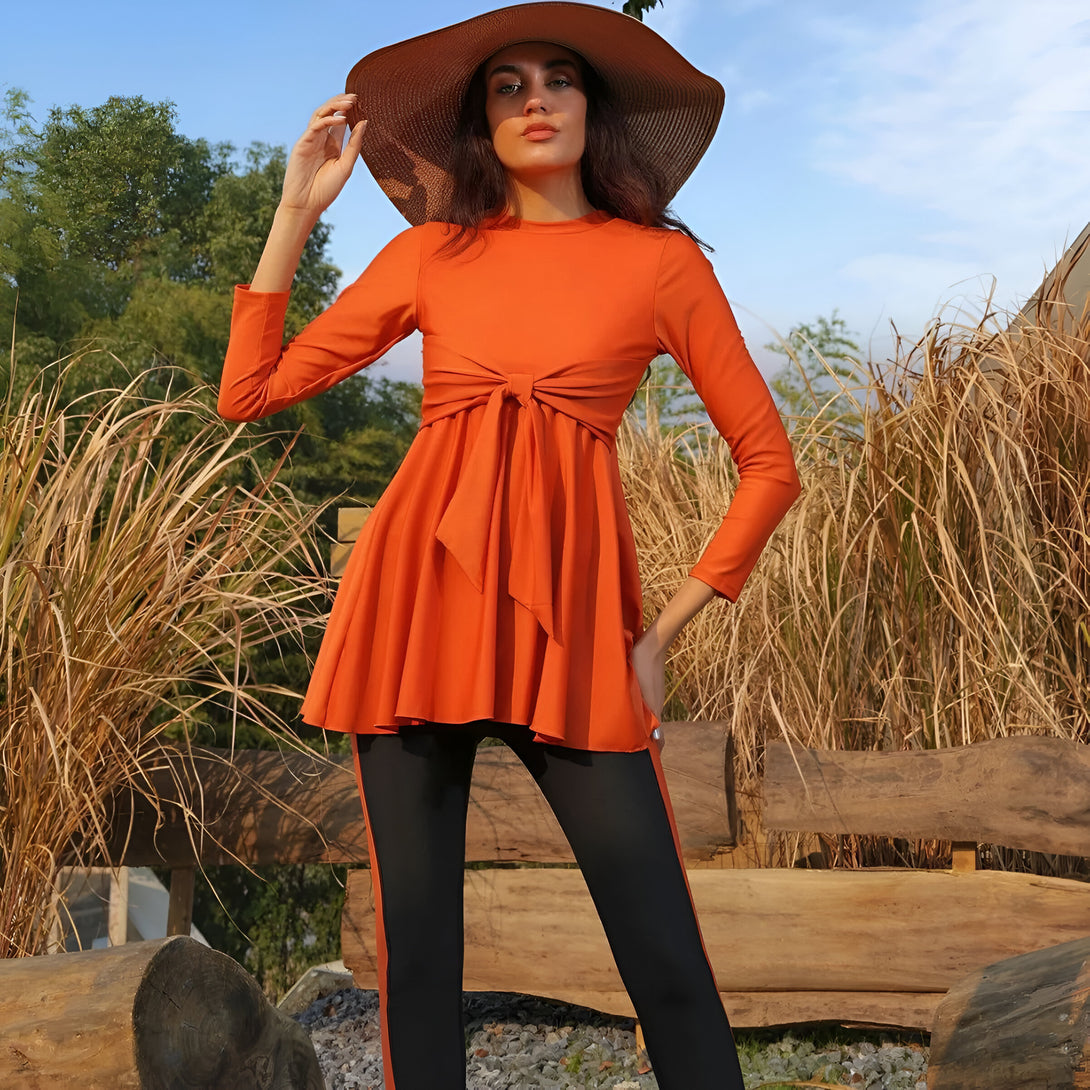 Une femme porte une robe orange à manches longues avec une ceinture nouée à la taille et un pantalon noir. Elle porte un chapeau de paille et est debout dans un champ d'herbe.