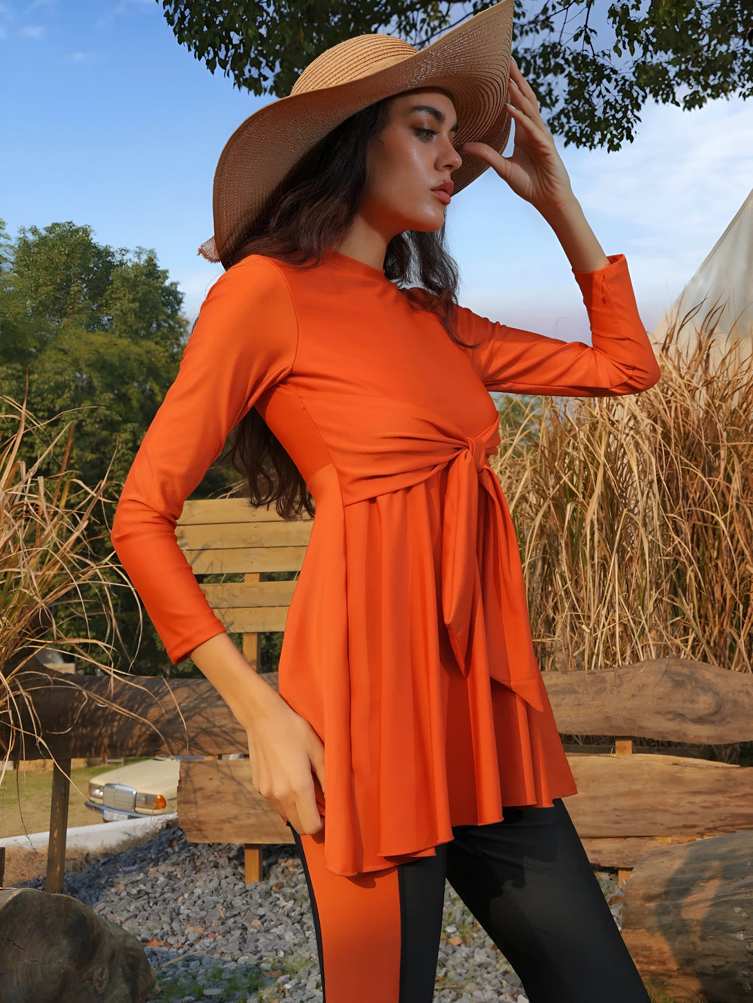 Une femme portant une robe longue orange avec une ceinture nouée à la taille, des leggings noirs et un chapeau de paille. Elle est debout dans un jardin.