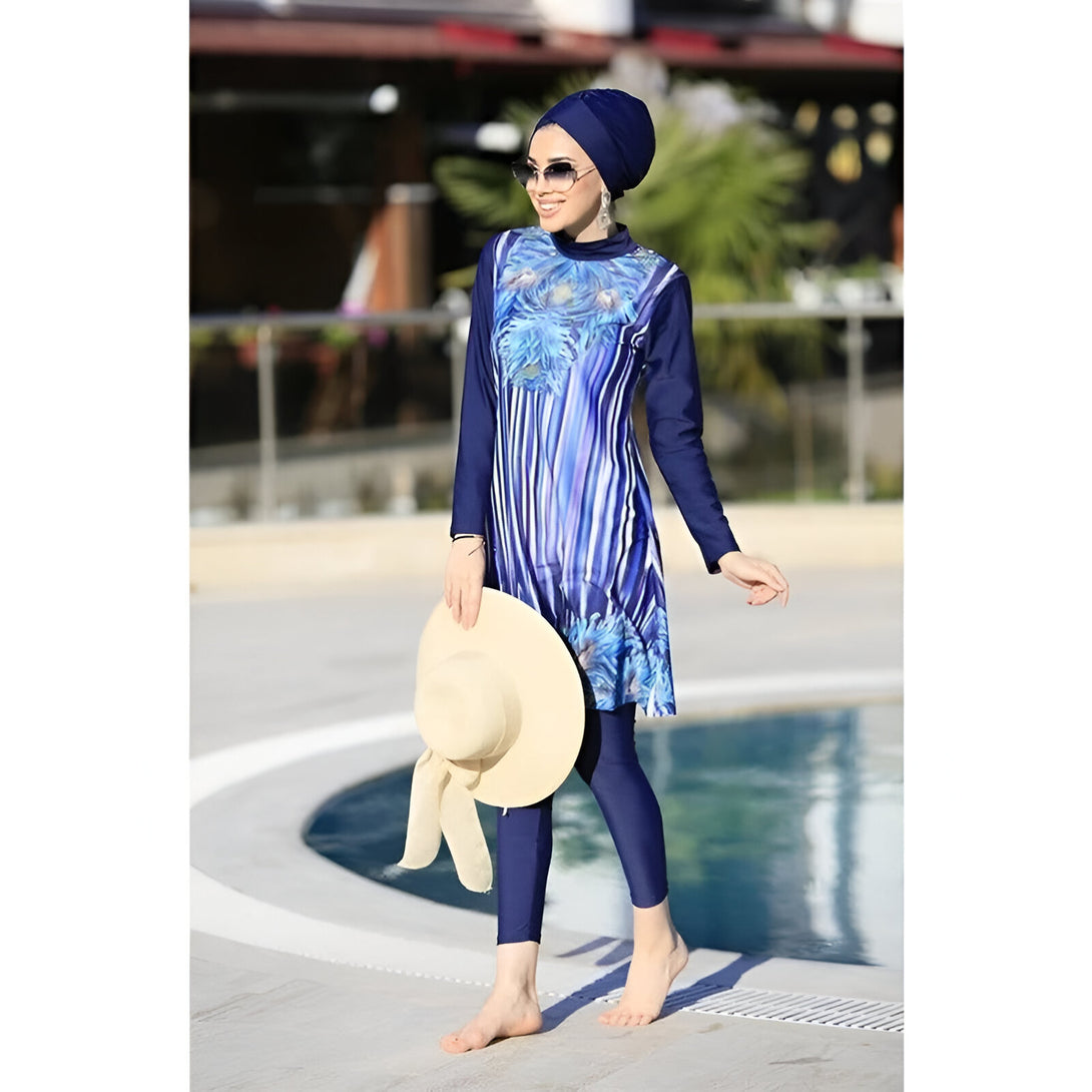 Une femme porte un maillot de bain musulman bleu marine à rayures avec un imprimé floral. Elle porte un hijab bleu marine et des lunettes de soleil. Elle est debout près d'une piscine et tient un chapeau de paille.