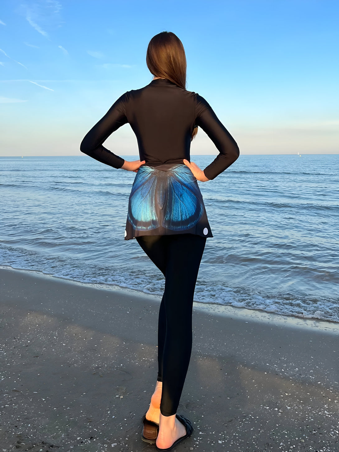 Une femme portant une tenue de plage musulmane noire avec un motif de papillon bleu sur la jupe, se tient sur une plage de sable et regarde l'océan.