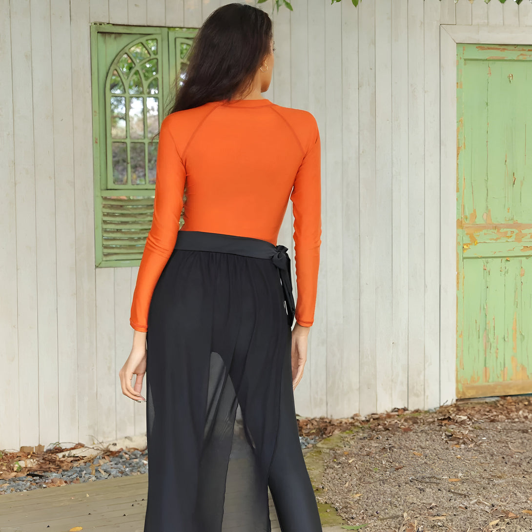 Une femme portant un haut orange à manches longues et une jupe noire transparente, avec une ceinture noire, de dos.