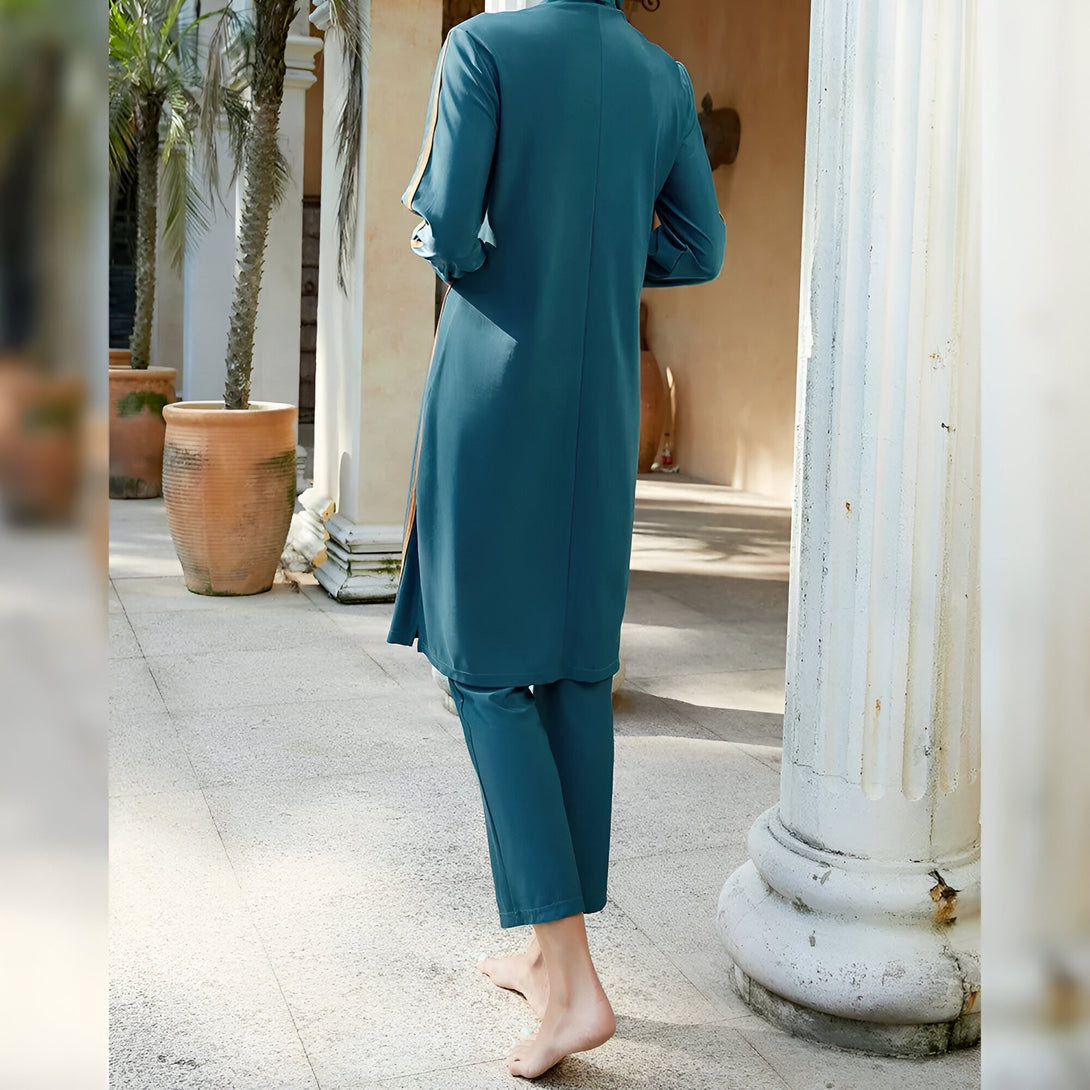 Une femme portant un maillot de bain musulman turquoise avec des détails dorés, debout devant une colonne de pierre.