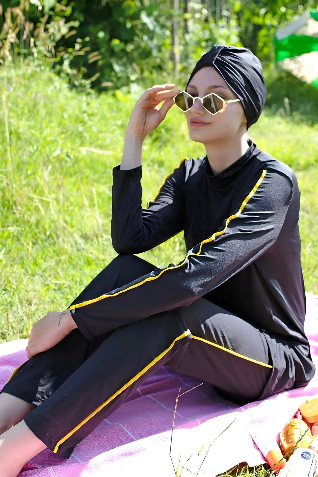 Une femme portant un maillot de bain musulman noir avec des détails jaunes, assise sur une serviette de plage rose.  Elle porte des lunettes de soleil et un foulard noir.  Parfait pour la natation ou la plage.