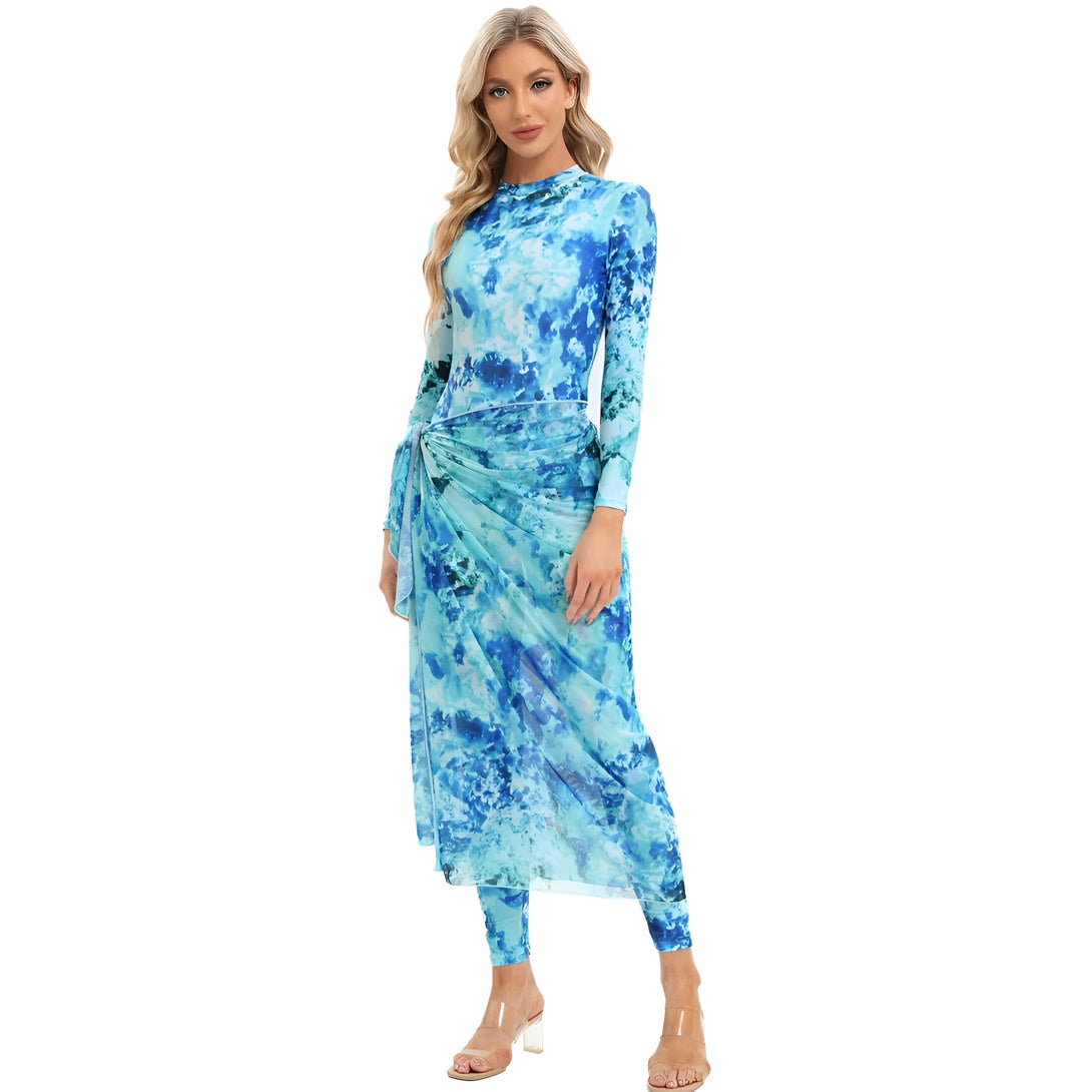 Une femme porte une robe longue bleue à manches longues avec un imprimé tie-dye et un tissu transparent. Elle porte également un pantalon long bleu sous la robe et des talons hauts transparents. La robe est nouée à la taille.