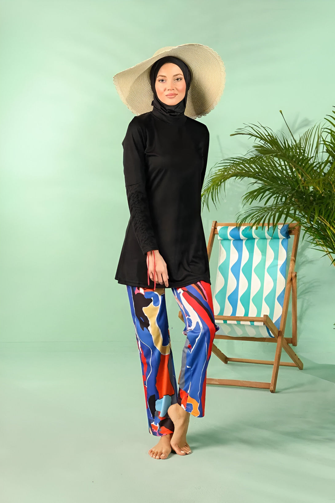 Femme portant un maillot de bain musulman noir avec des manches longues et un pantalon coloré, tenant un chapeau de paille.