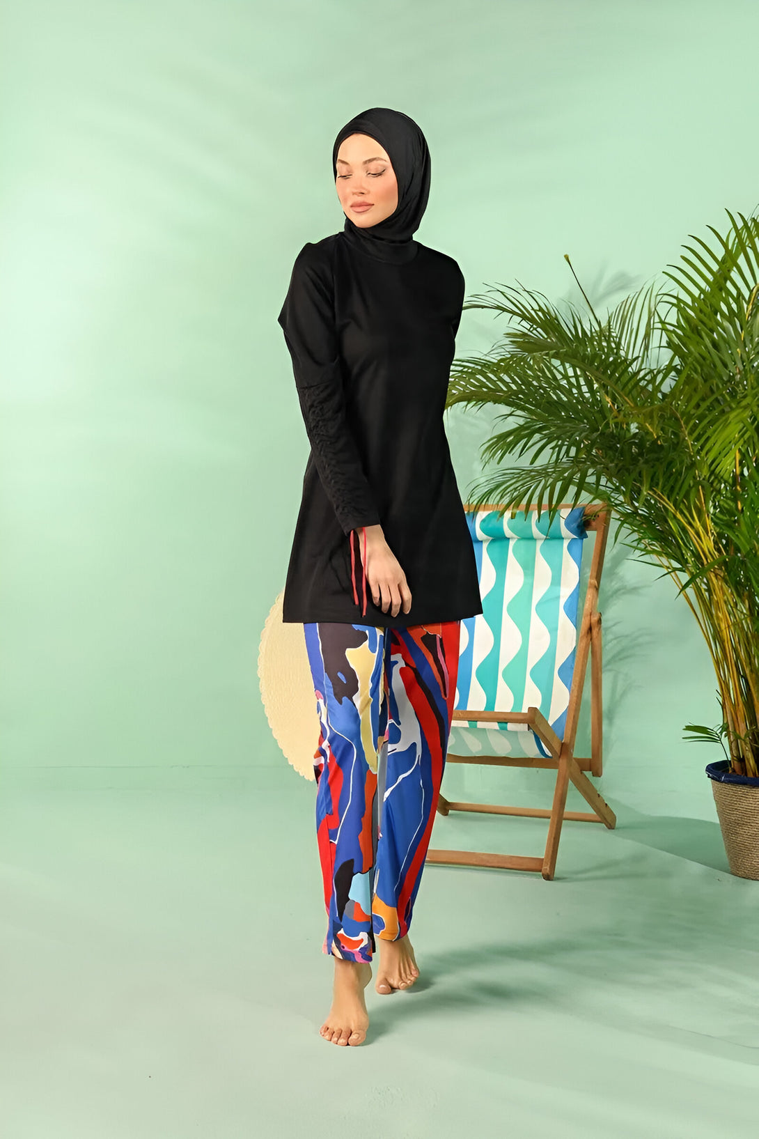 Une femme portant un hijab et un maillot de bain musulman noir avec un pantalon coloré. Elle est dans une ambiance estivale avec un fond vert et une chaise de plage en bois.