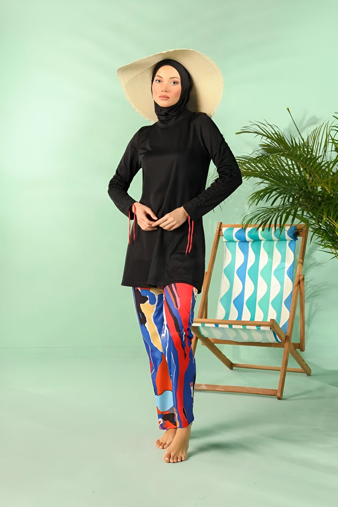 Une femme portant un maillot de bain musulman noir avec des détails rouges et un pantalon coloré, debout devant une chaise de plage sur un fond vert.