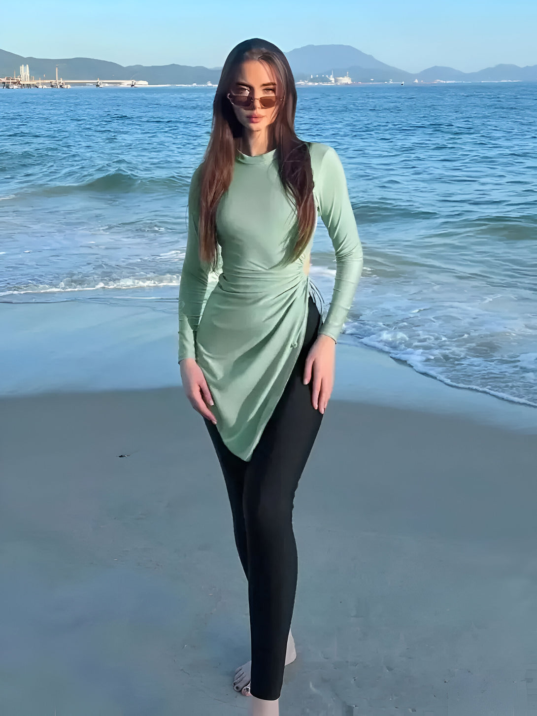 Une femme portant un hijab vert et des lunettes de soleil se tient sur une plage. Elle porte un pantalon noir et un haut vert qui couvre tout son corps. La tenue est parfaite pour aller à la plage et rester modeste.