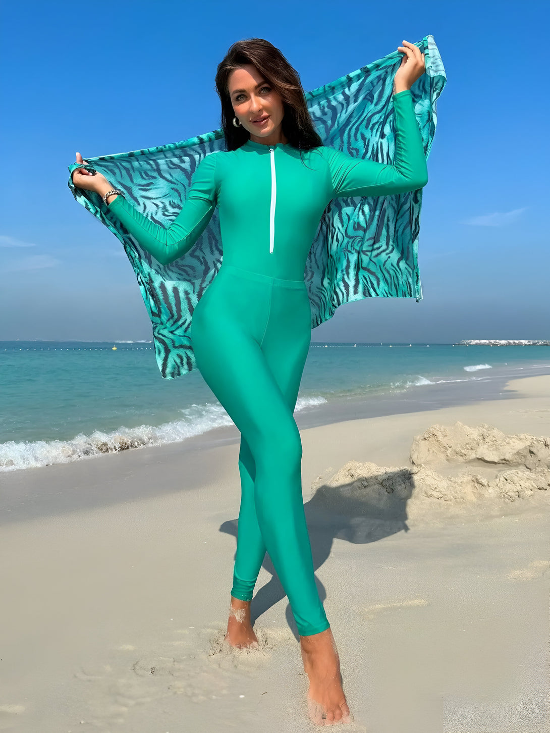 Une femme portant un maillot de bain vert à manches longues et un foulard à imprimé animal sur la plage.