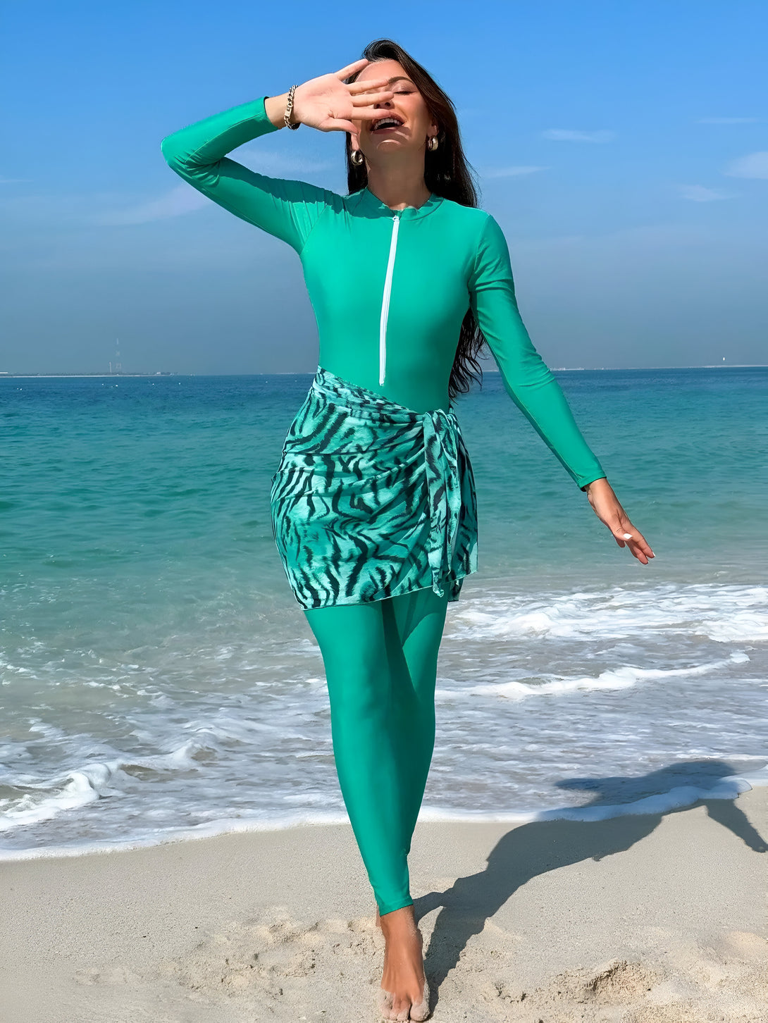 Une femme souriante porte un maillot de bain islamique vert sur la plage. Le maillot est assorti d'une jupe en tissu imprimé zèbre. Elle est pieds nus sur le sable blanc.