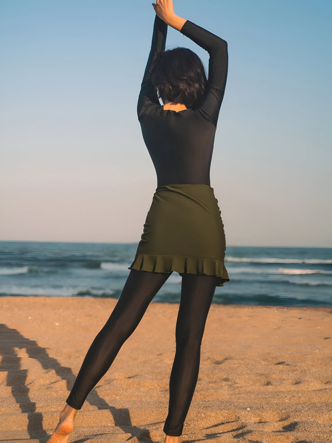 Une femme portant une tenue de plage musulmane élégante et confortable, avec un haut noir à manches longues et une jupe verte à volants, se tient sur la plage face à la mer.
