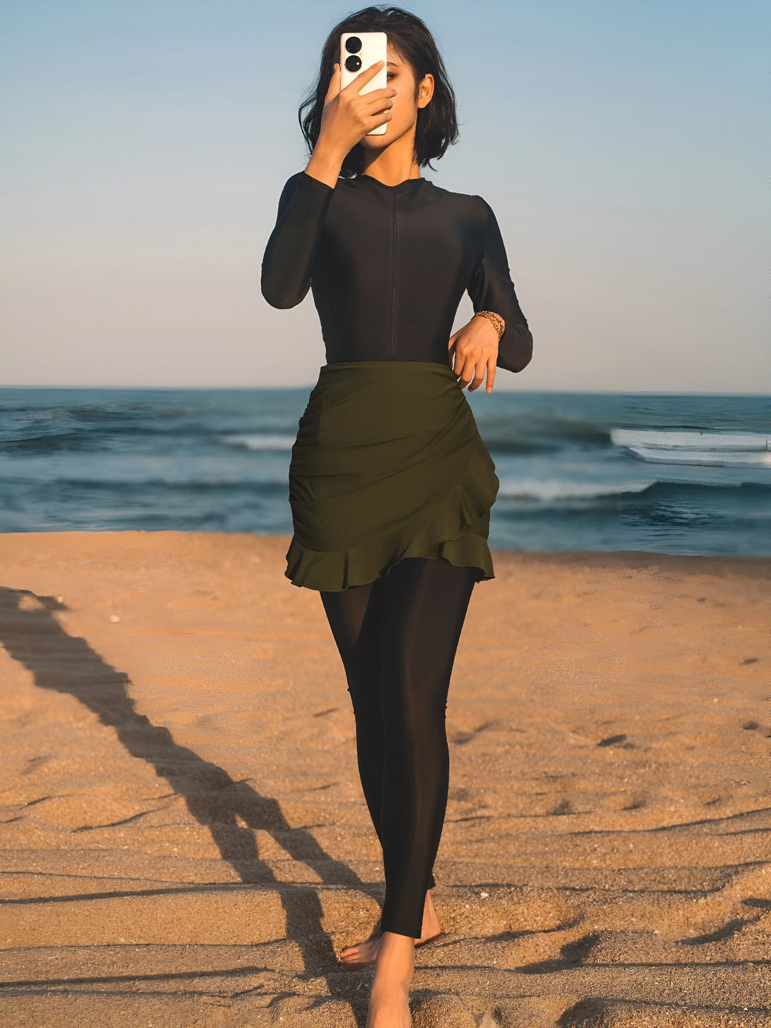 Une femme portant un maillot de bain musulman vert et noir se promène sur la plage.