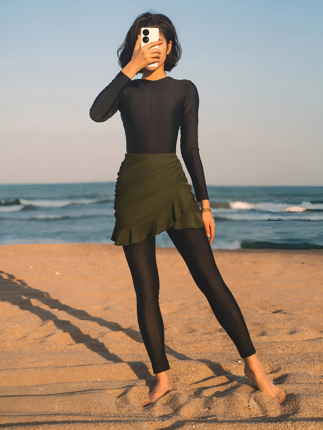 Une femme portant un maillot de bain musulman noir et une jupe verte se tient sur la plage.  Elle regarde son téléphone.  Parfait pour la plage ou la piscine !