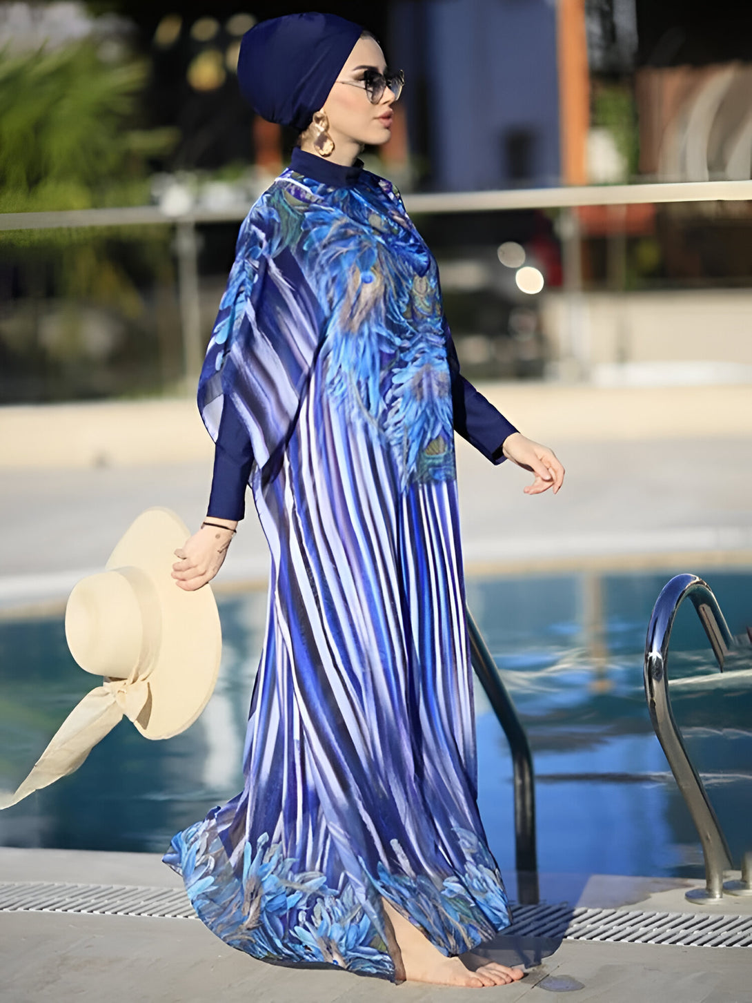 Une femme portant un hijab bleu marine et une robe de bain islamique à rayures bleues et blanches avec un motif floral près d'une piscine.