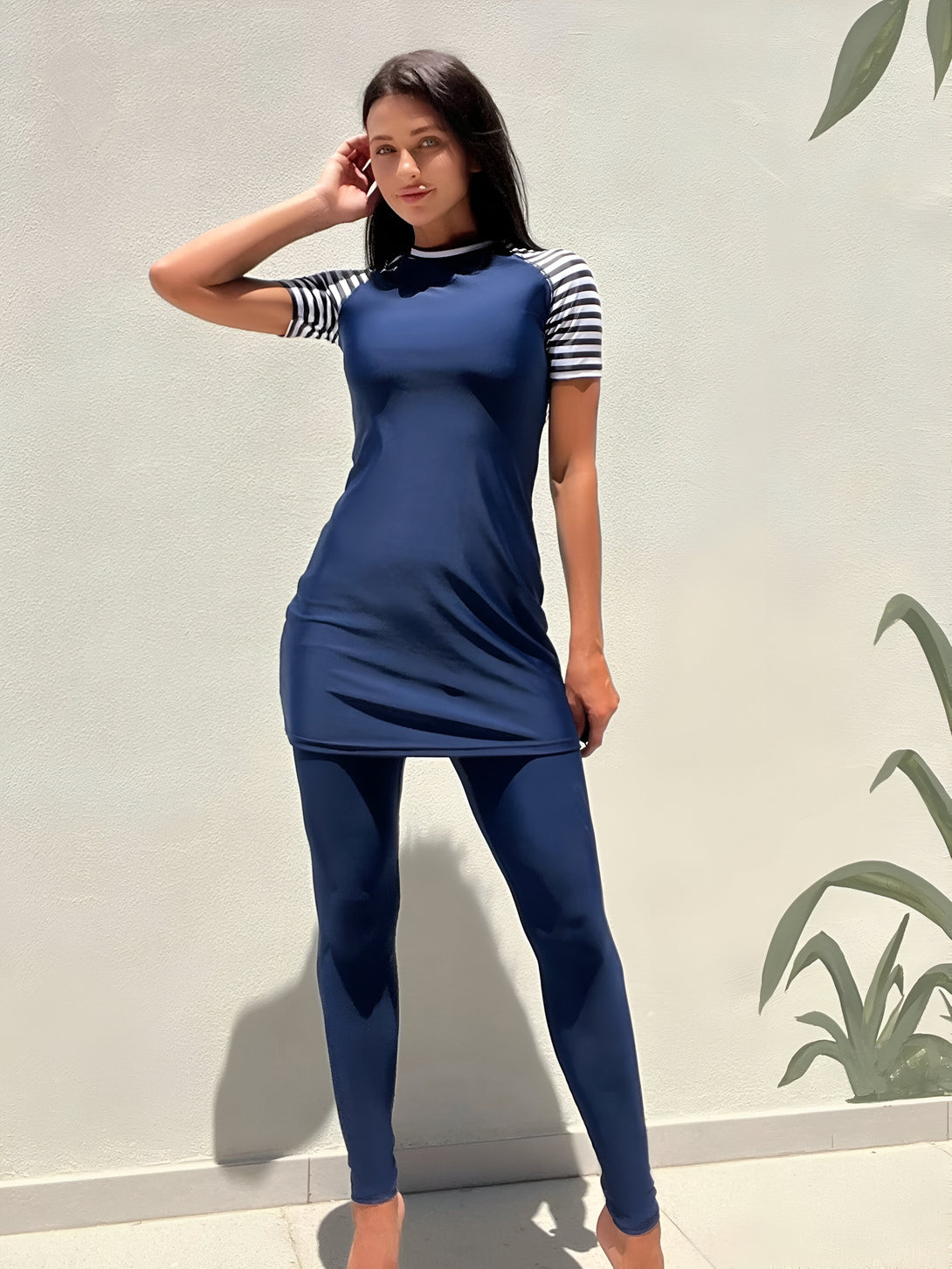 Une femme portant un maillot de bain musulman bleu marine avec des manches rayées blanches et noires.  Le maillot de bain est composé d'une robe longue et d'un legging.  La femme est debout devant un mur blanc.