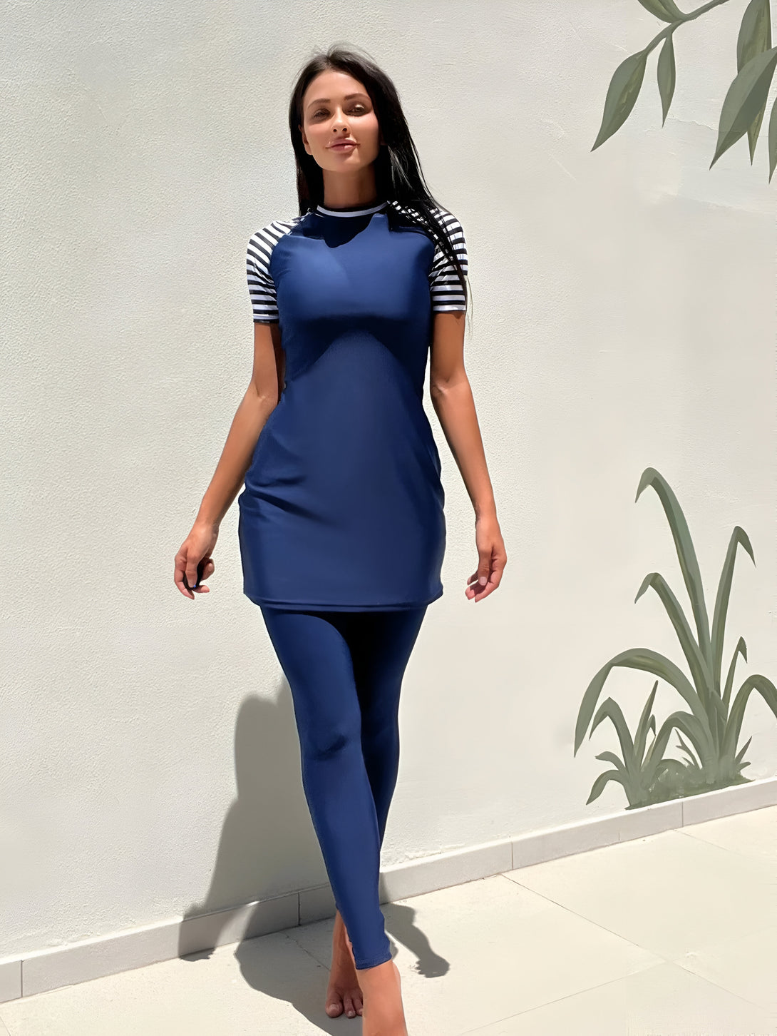 Une femme portant un maillot de bain musulman bleu marine avec des manches courtes rayées blanches et noires. Le maillot de bain est composé d'une robe et d'un pantalon. La femme se tient devant un mur blanc avec des plantes vertes en arrière-plan.