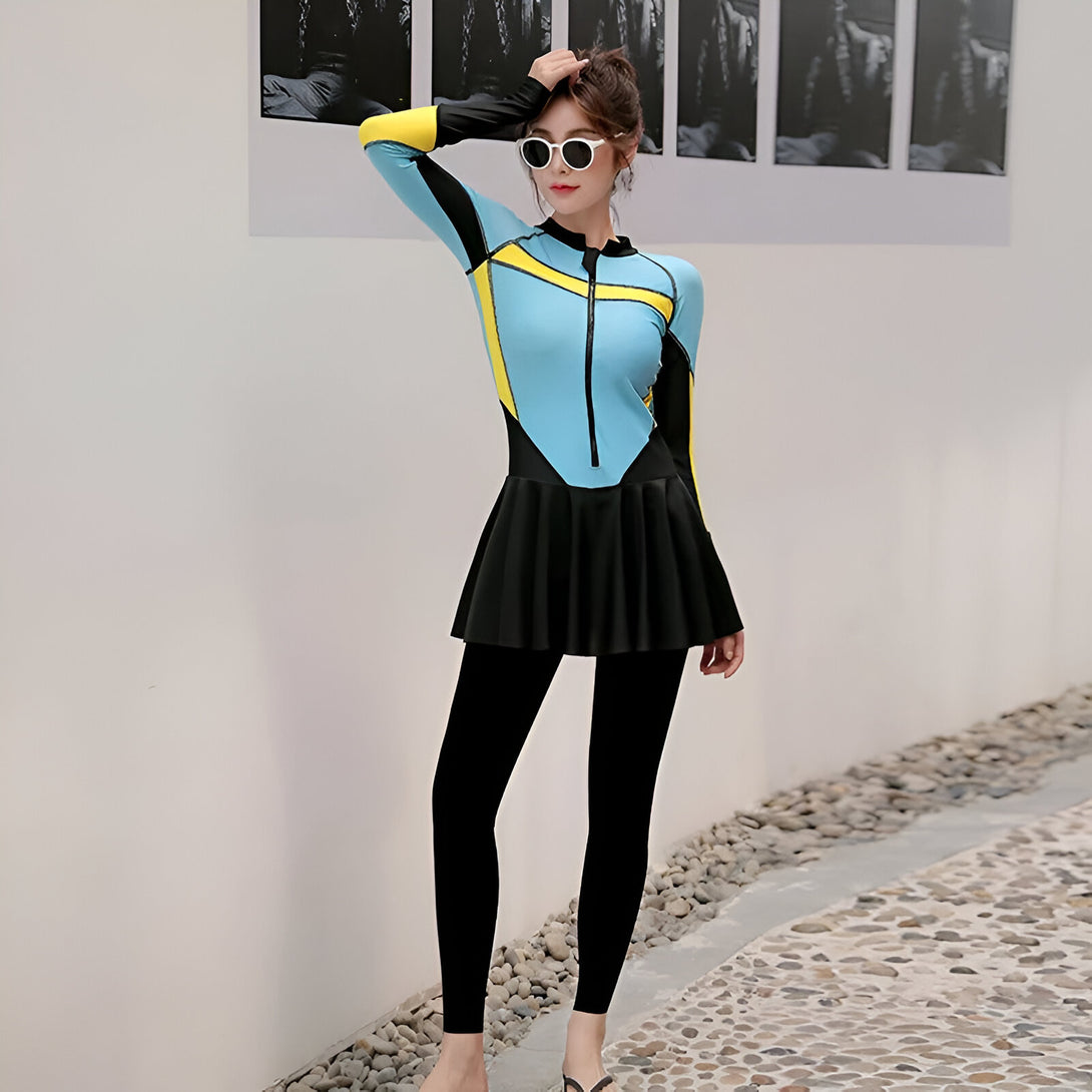 Une femme portant un maillot de bain musulman bleu et jaune avec des leggings noirs. Le maillot de bain a une jupe intégrée et des manches longues. La femme porte des lunettes de soleil et sourit à la caméra.