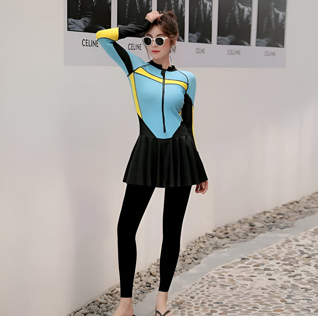 Une femme porte un maillot de bain musulman bleu, jaune et noir avec une jupe et des leggings. Elle porte également des lunettes de soleil et sourit.