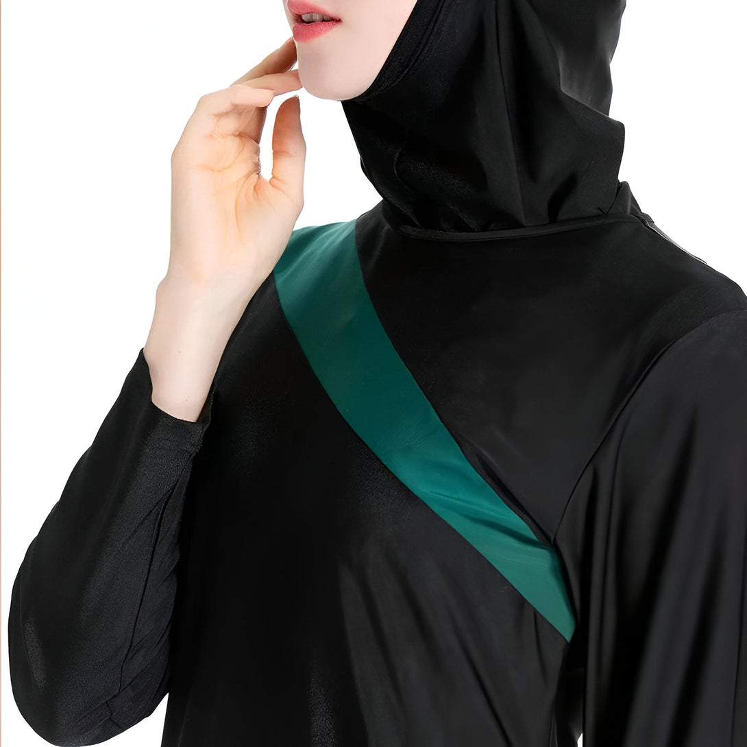 Une femme porte un maillot de bain musulman noir avec une bande verte diagonale.  Le maillot de bain a des manches longues et une capuche.