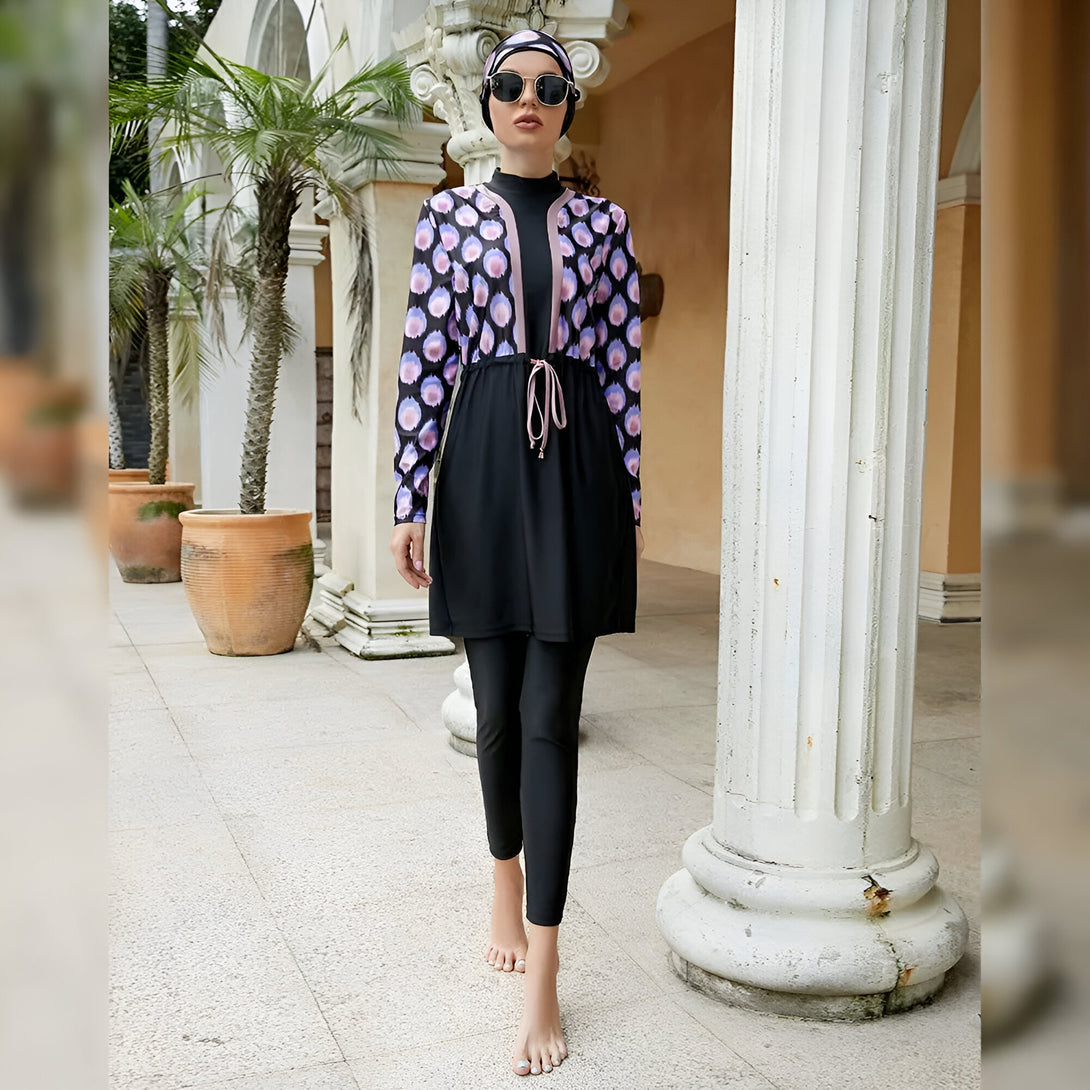 Une femme portant un maillot de bain musulman à manches longues avec un motif floral. Le maillot de bain est noir avec des accents de couleur rose et est assorti d'un hijab assorti. La femme porte également des lunettes de soleil et sourit à la caméra. Elle marche sur une allée pavée.