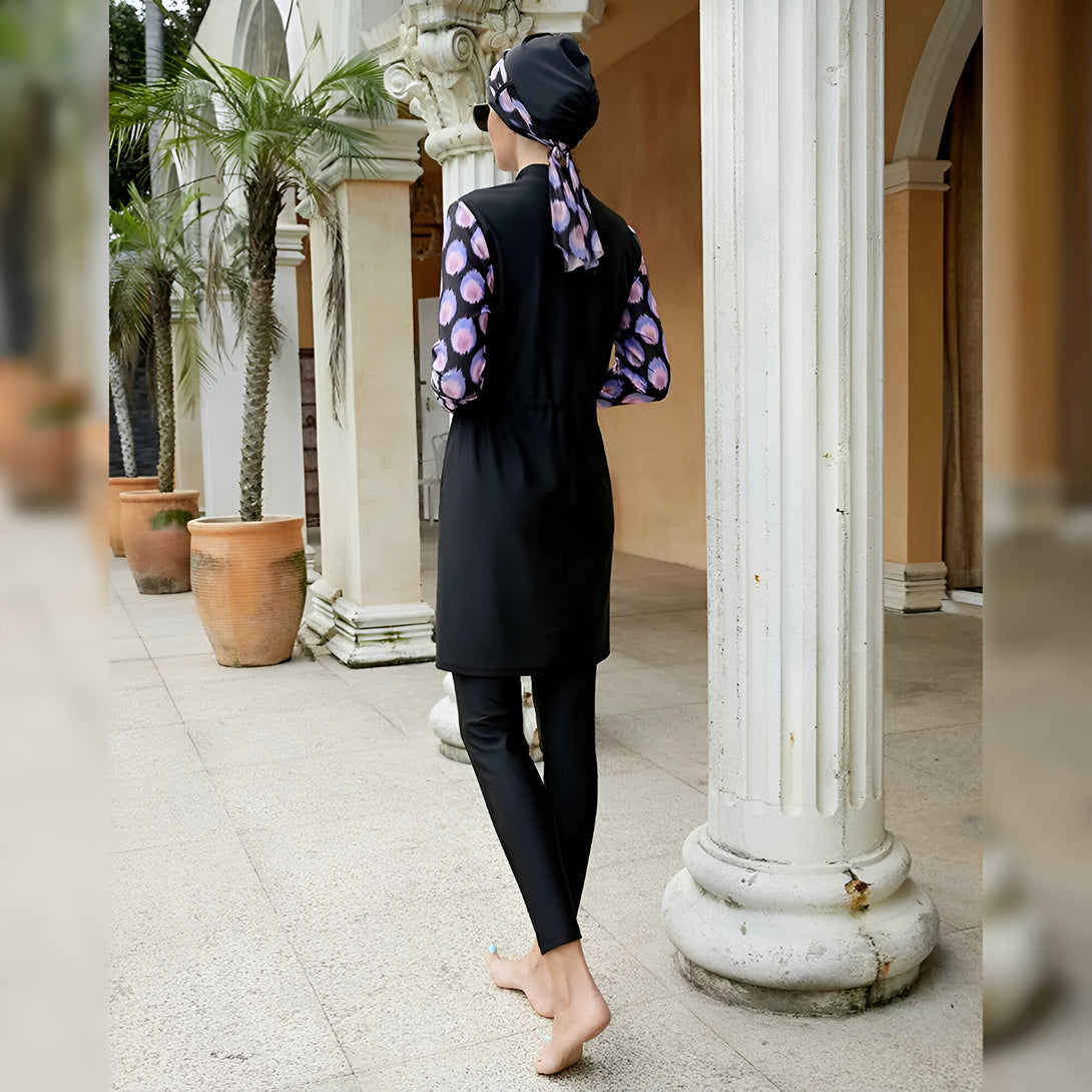 Une femme portant un maillot de bain musulman noir avec une tunique et un pantalon. La tunique est décorée de motifs floraux colorés et elle porte un hijab assorti. Elle se tient dans un jardin avec des colonnes blanches.