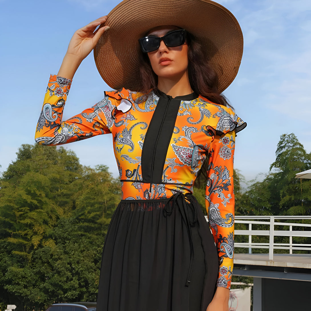 Une femme portant un maillot de bain musulman à manches longues avec un motif floral orange et bleu. Elle porte un chapeau de paille et des lunettes de soleil.
