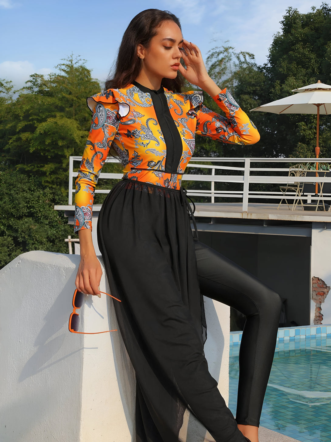 Une femme portant un maillot de bain musulman orange à motifs et une jupe noire transparente près d'une piscine.