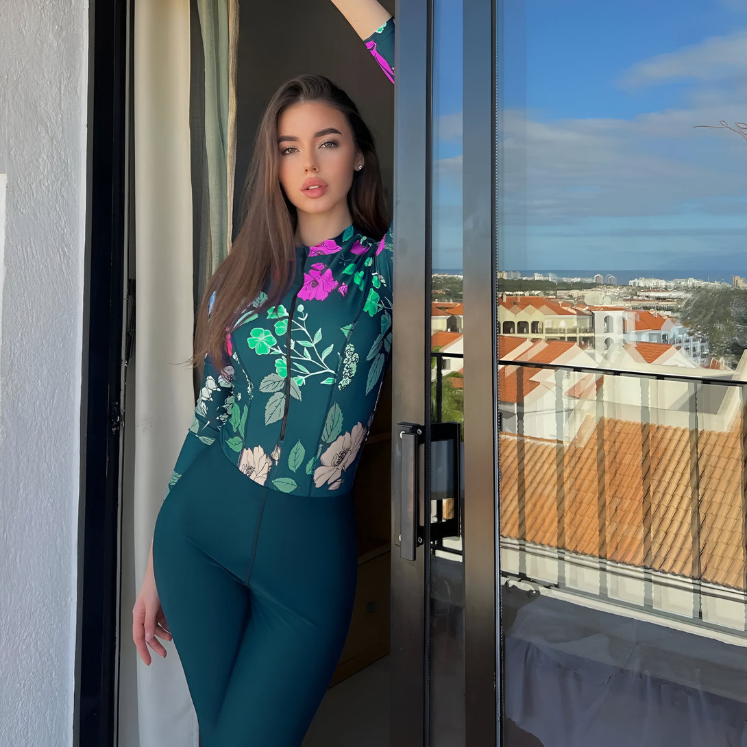 Une femme portant un ensemble de vêtements musulmans vert avec des fleurs. Elle se tient près d'une porte-fenêtre avec une vue sur la mer.