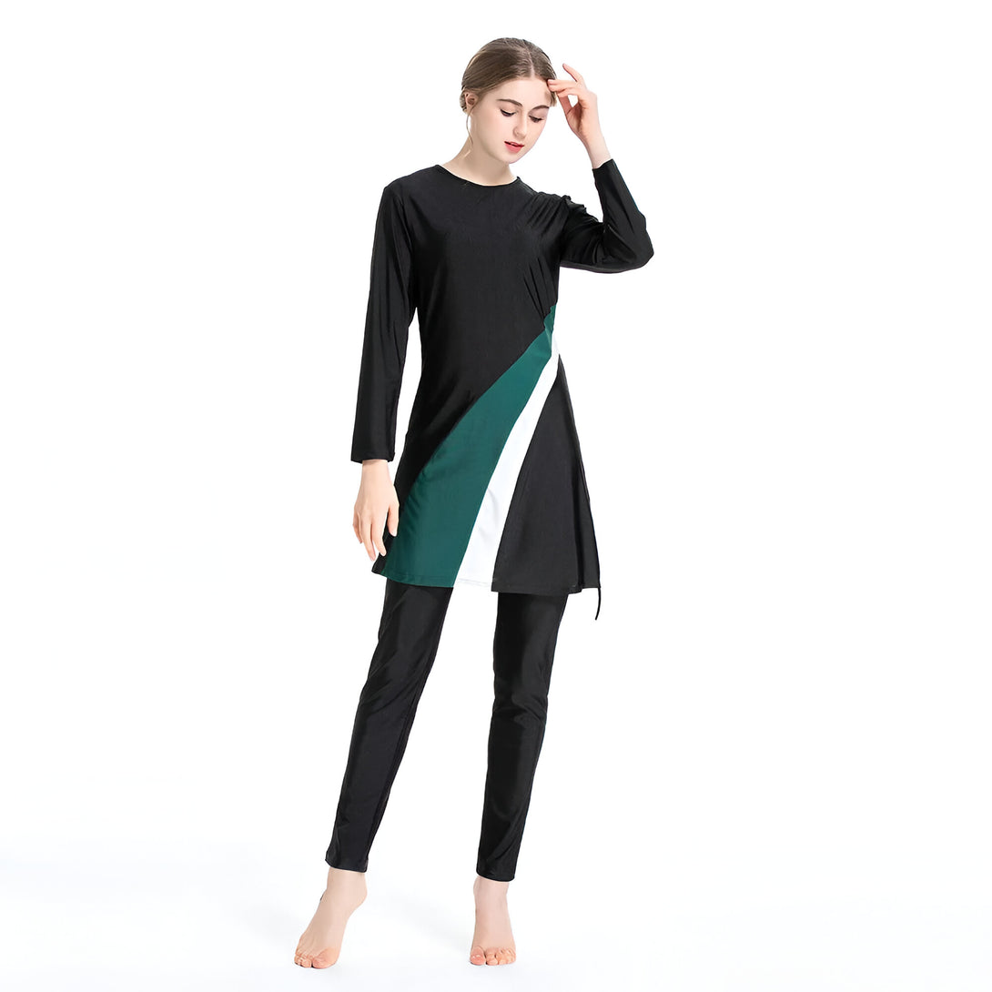 Une femme porte un maillot de bain musulman noir avec des détails verts et blancs. Le maillot de bain a des manches longues et une jupe longue.  La femme est debout sur un fond blanc.