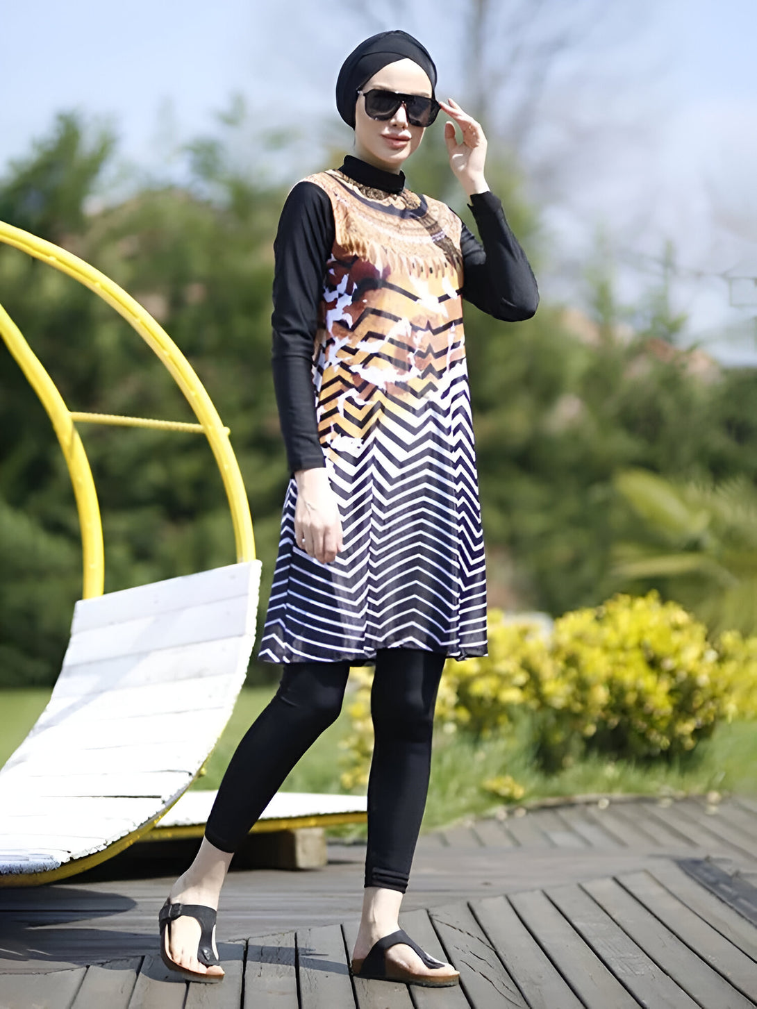 Une femme portant un maillot de bain musulman noir et blanc à rayures avec un imprimé géométrique doré. Elle porte également un hijab noir et des lunettes de soleil.