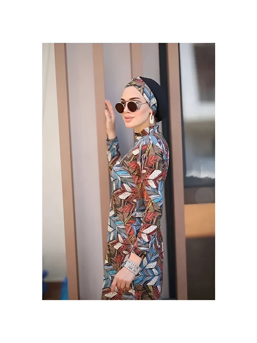 Une femme portant un hijab noir et une robe de bain à manches longues avec un motif coloré. Elle porte des lunettes de soleil rondes.