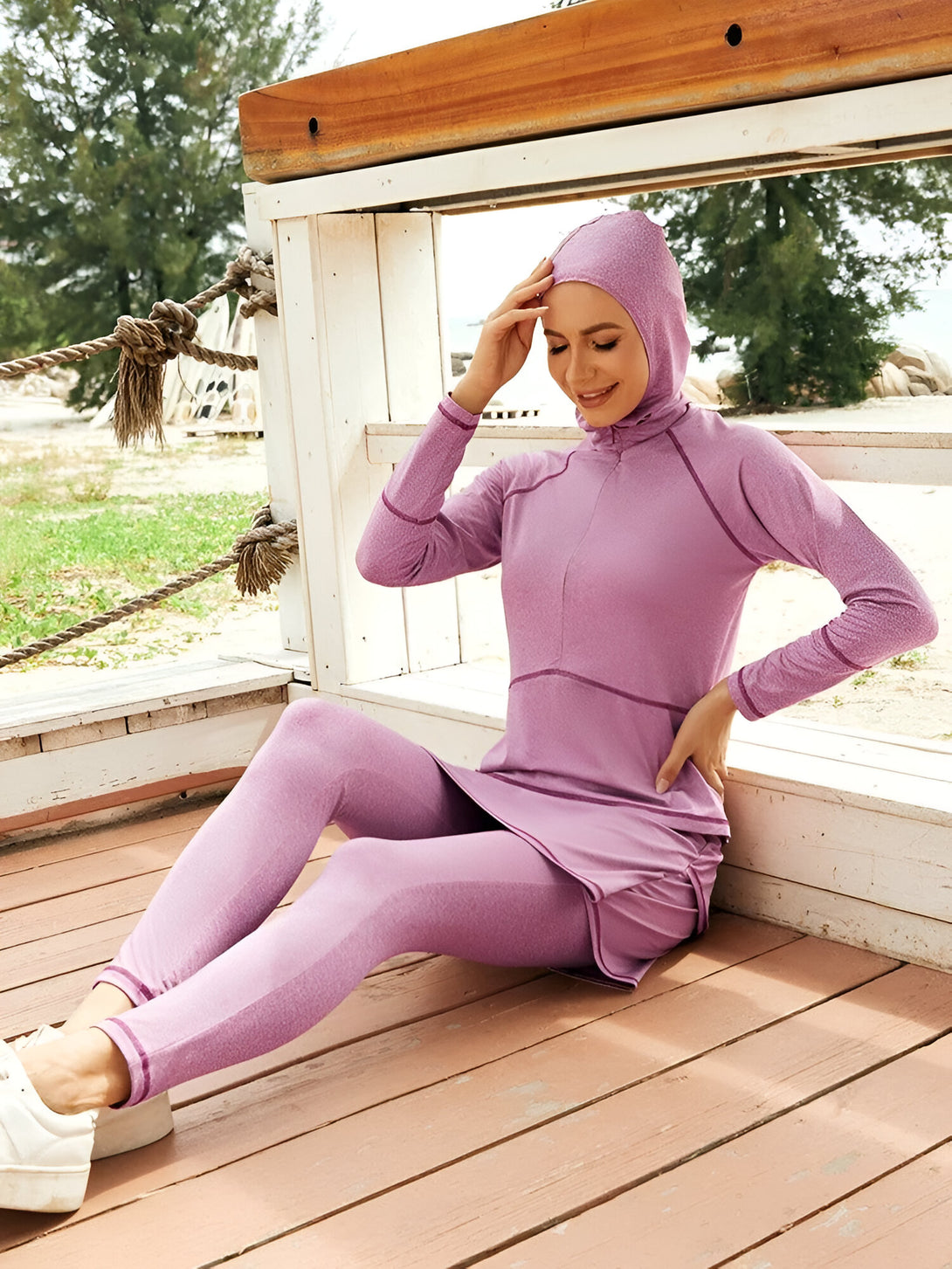 Une femme porte un ensemble de vêtements de bain musulmans de couleur lilas, composé d'un haut à manches longues, d'un hijab assorti et d'un short de plage. Elle est assise sur un pont en bois.