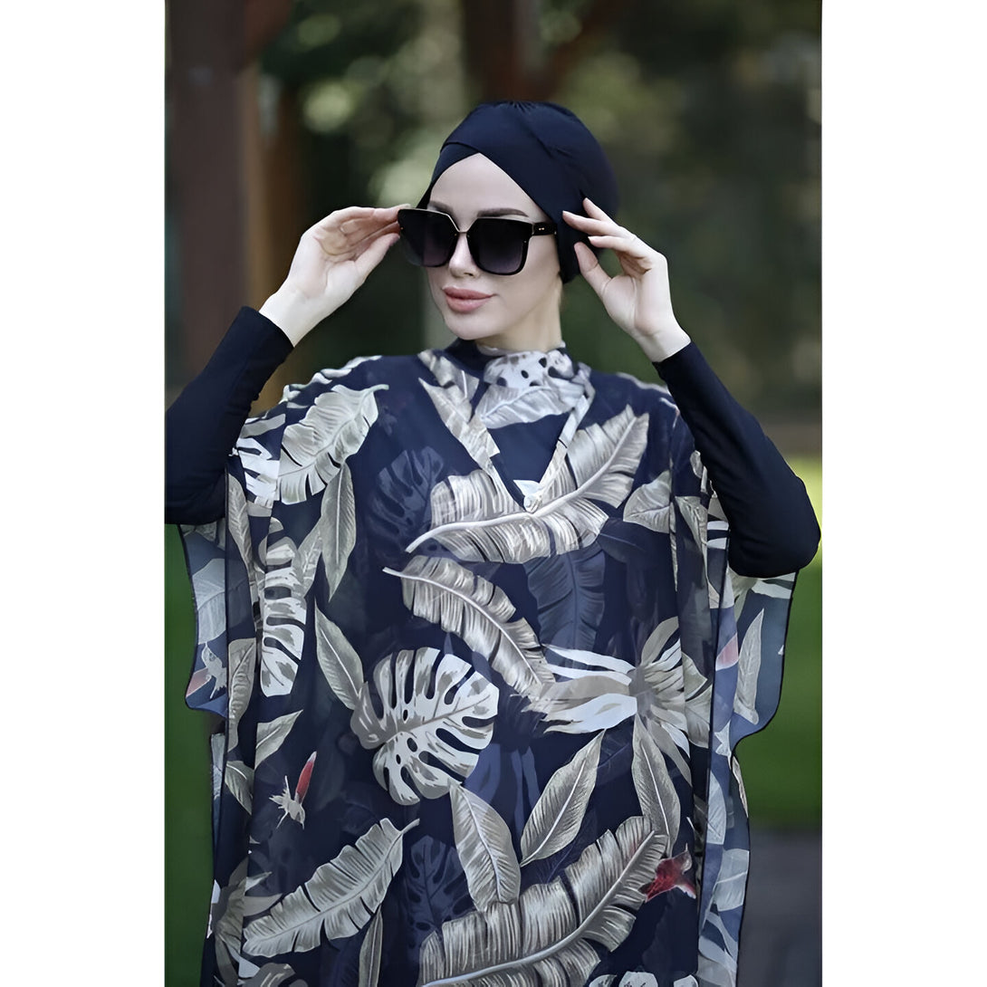 Une femme portant un hijab noir et un tunique de plage à imprimé tropical.  Le tunique est bleu foncé avec des feuilles blanches et beige.