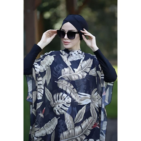Une femme portant un hijab noir et un tunique de plage à imprimé tropical.  Le tunique est bleu foncé avec des feuilles blanches et beige.