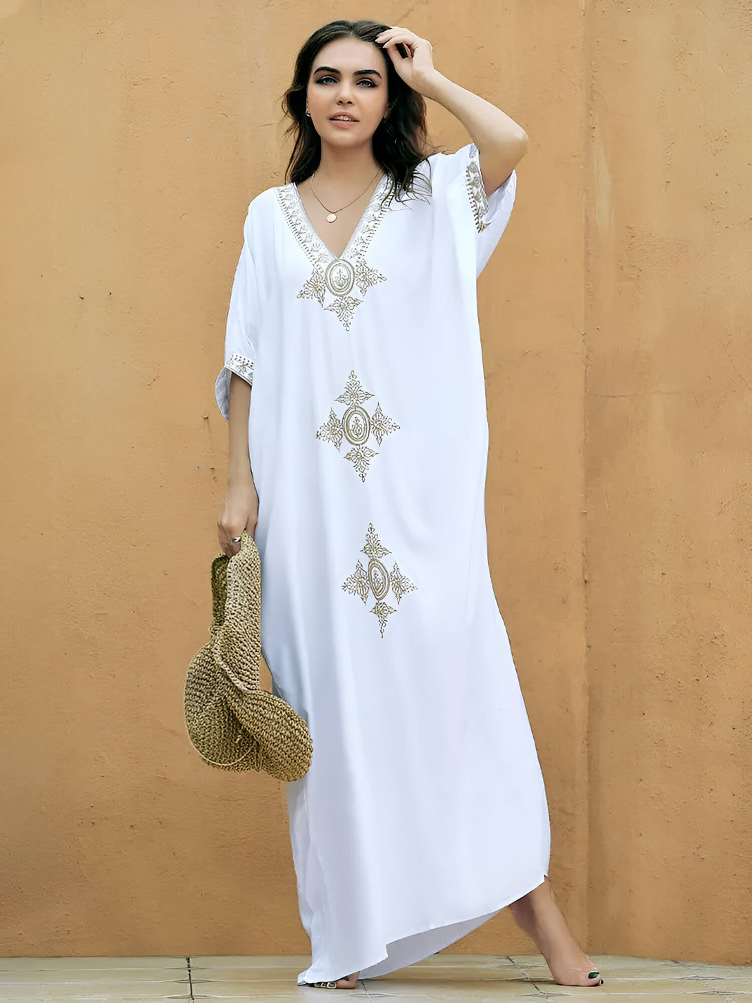 Une femme porte une longue robe blanche avec des broderies dorées. La robe a un col en V et des manches courtes. La femme porte un sac à main en paille et sourit à la caméra.