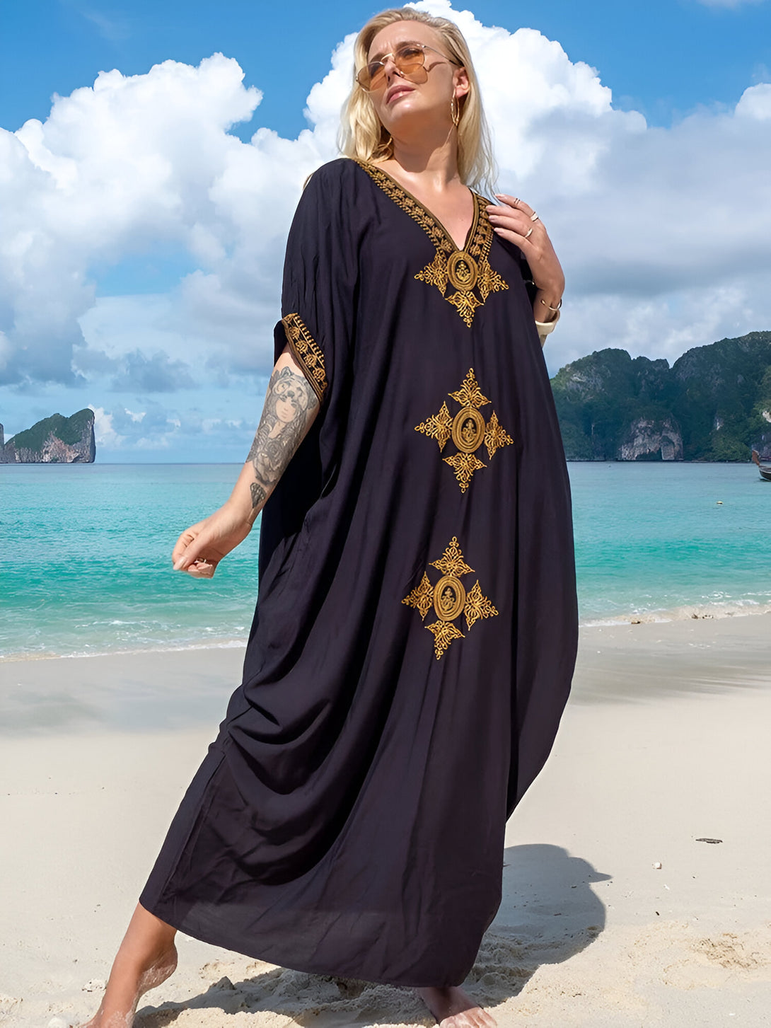 Une femme portant une longue robe noire à manches courtes et des broderies dorées, se tient sur une plage de sable fin, sous un ciel bleu azur.