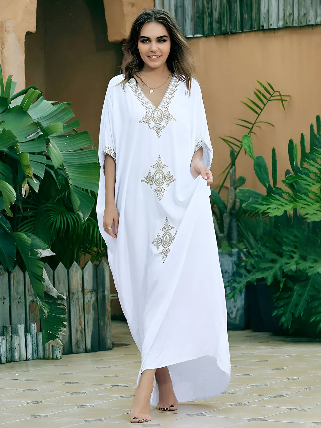 Une femme souriante porte une longue robe blanche avec des motifs dorés. La robe est à manches courtes et a un col en V. La femme est debout dans un jardin.