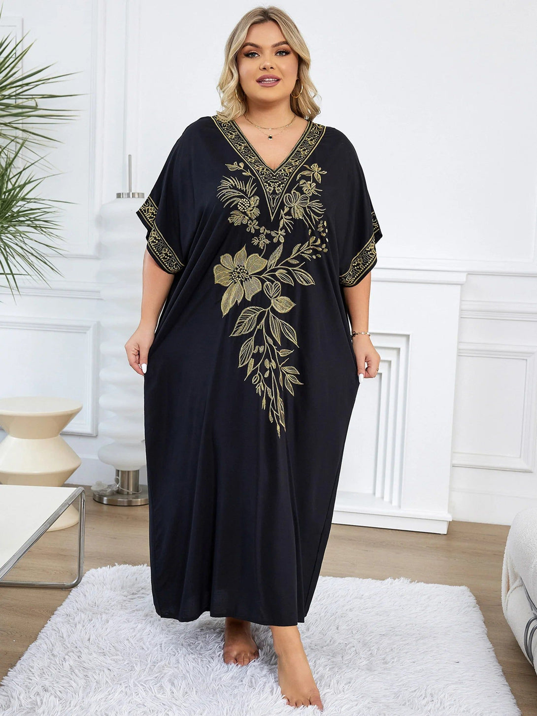 Une femme porte une longue robe noire avec un motif floral doré. La robe a une encolure en V et des manches courtes. La femme est debout sur un tapis blanc.