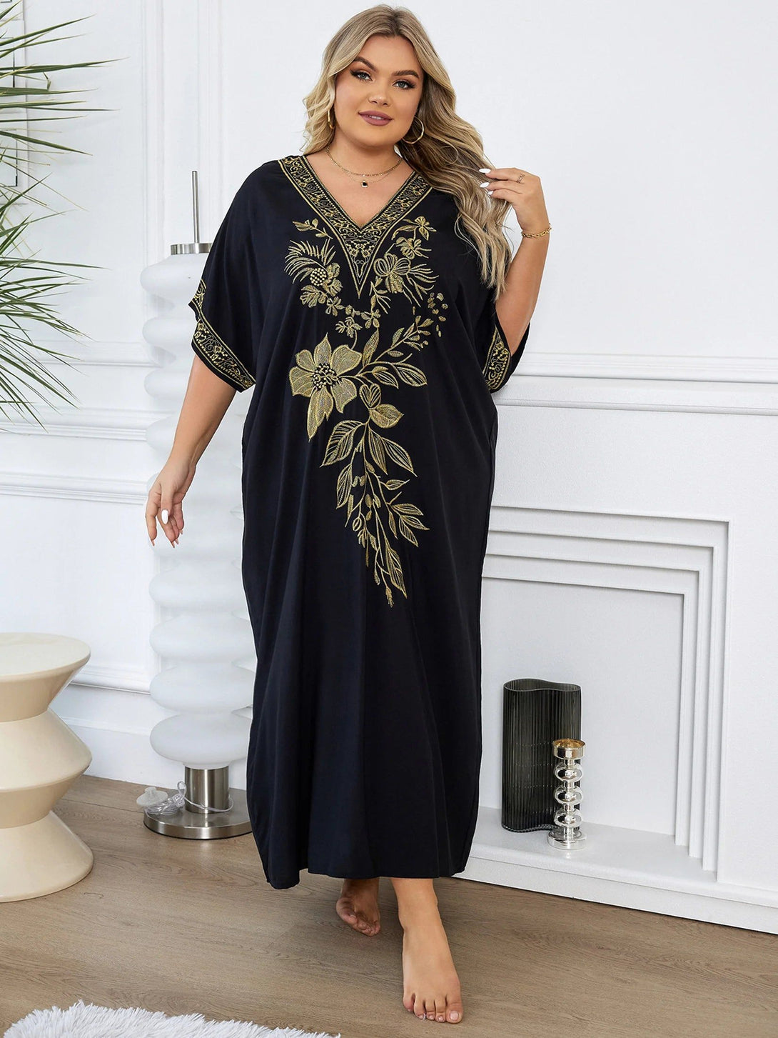 Une femme porte une robe noire longue avec des motifs floraux dorés. La robe a une encolure en V et des manches courtes.