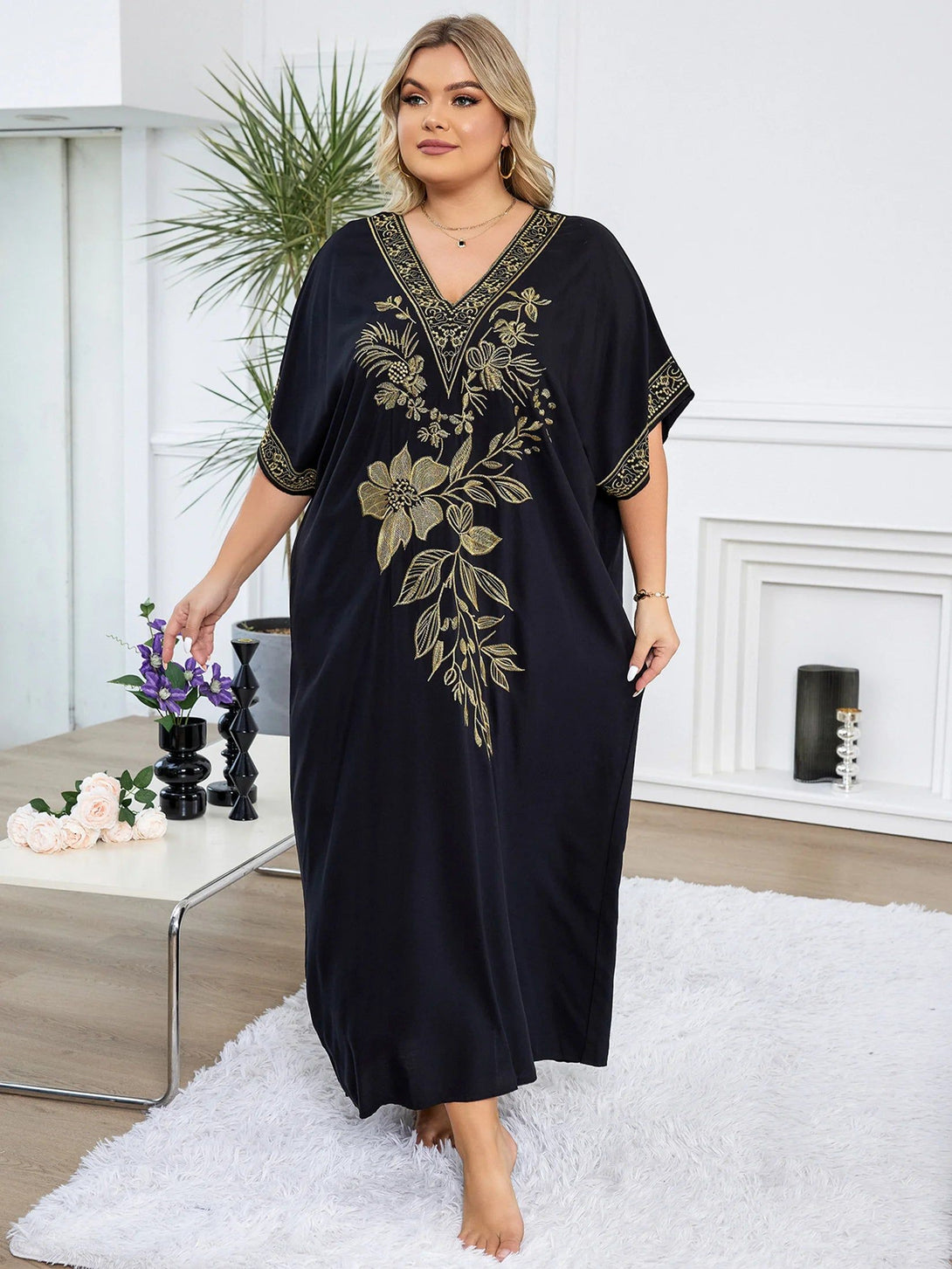 Une femme portant une longue robe noire à manches courtes avec un motif floral doré. La robe a un col en V et est ample. La femme est debout sur un tapis blanc moelleux.