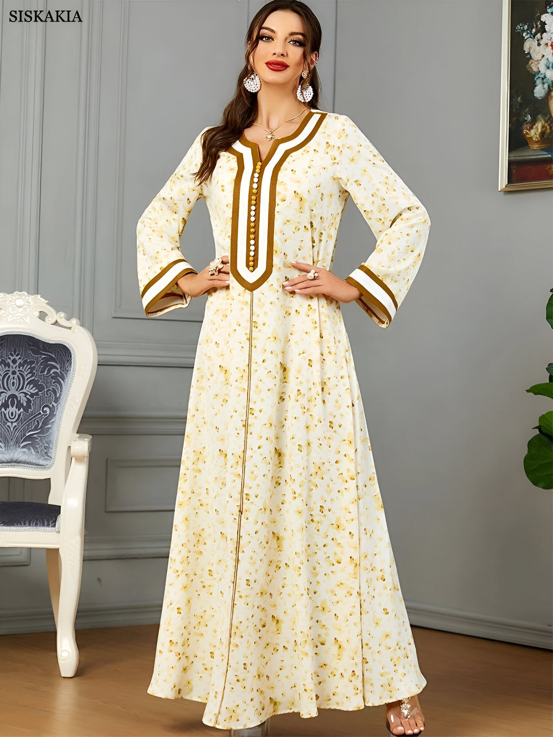 Une femme portant une longue robe blanche à fleurs avec des détails dorés. La robe a un col en V et des manches longues. La femme porte des boucles d'oreilles et un collier.