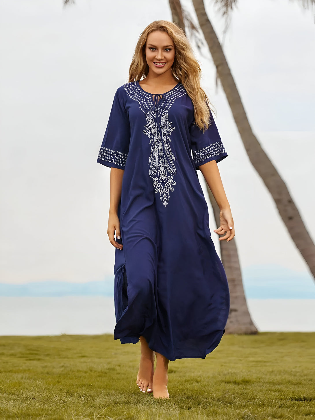 Une femme souriante porte une robe longue bleue marine avec des broderies blanches sur une plage. La robe est élégante et confortable, idéale pour les occasions spéciales ou les sorties décontractées.