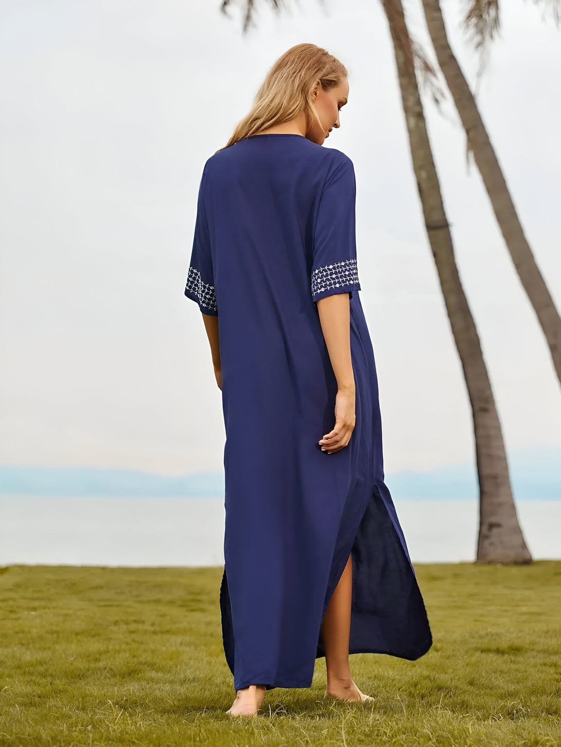 Une femme portant une longue robe bleue avec des détails brodés blancs, debout sur l'herbe devant un palmier.