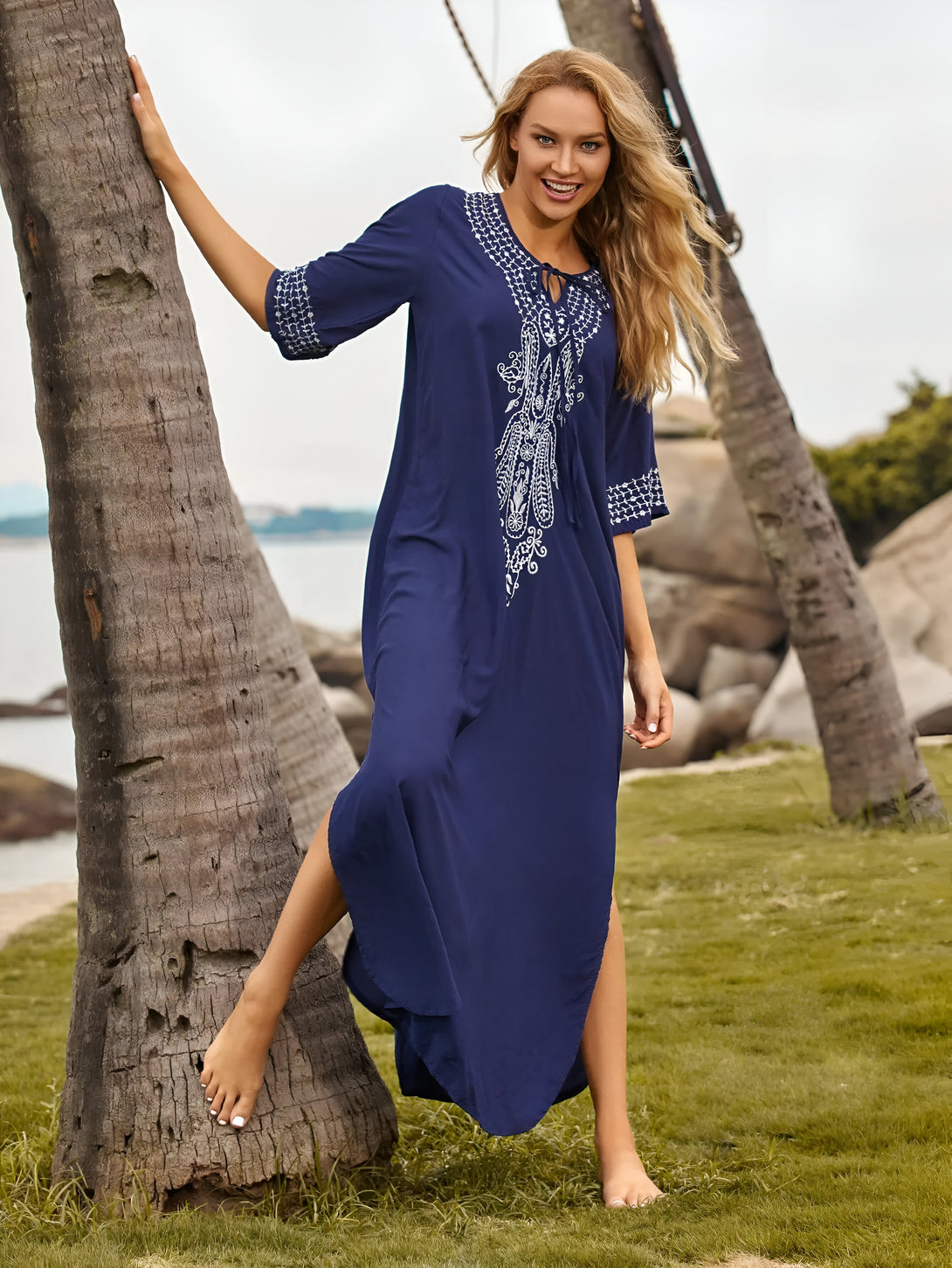 Une femme souriante porte une longue robe bleue en coton avec des détails brodés blancs. La robe est ample et fluide, idéale pour les journées chaudes. La femme est debout près d'un palmier sur une plage.